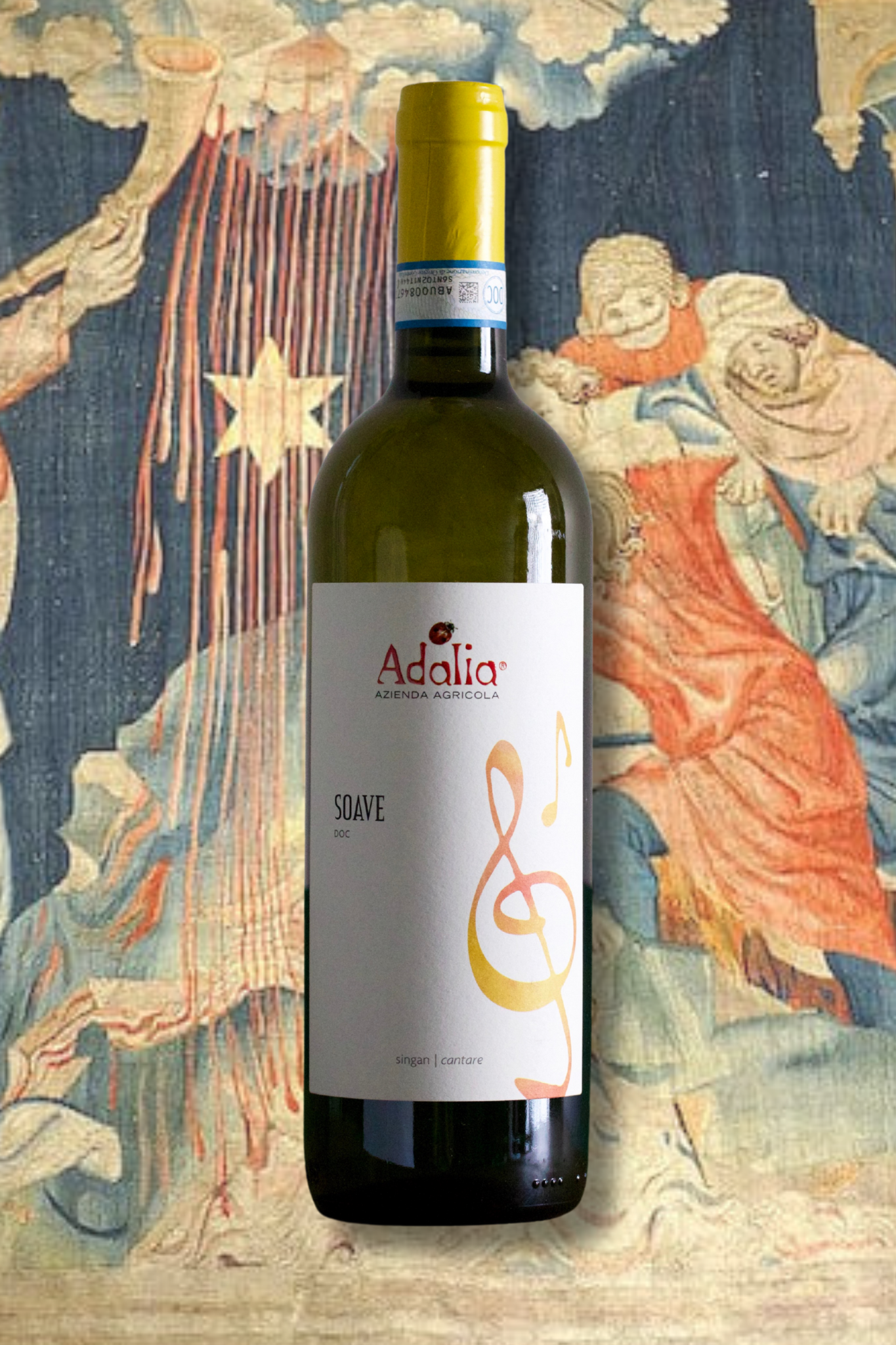 Adalia 'Singan' Soave 2023 bottle in front of a medieval-style tapestry featuring angels and musical motifs. Organic Garganega white wine from Veneto, Italy.
